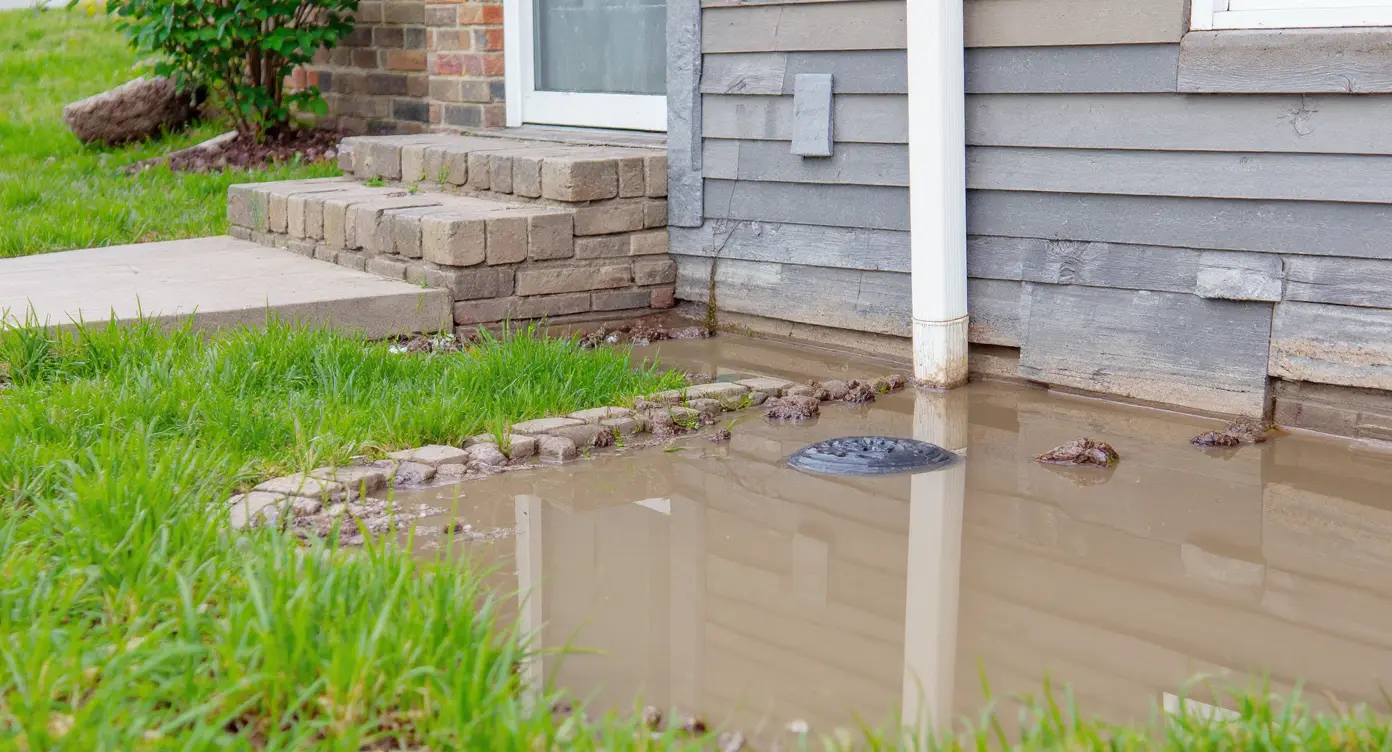 Exterior drainage failure showing downspout discharge too close to foundation with poor grading causing water pooling at the foundation wall base