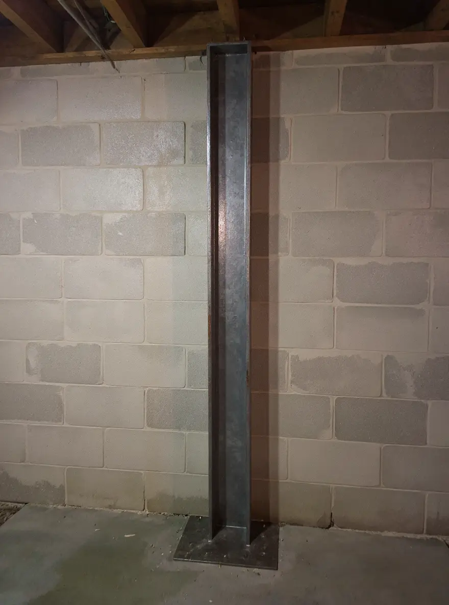 Vertical steel I-beam brace installed against a bowing basement wall, anchored to floor and ceiling joists